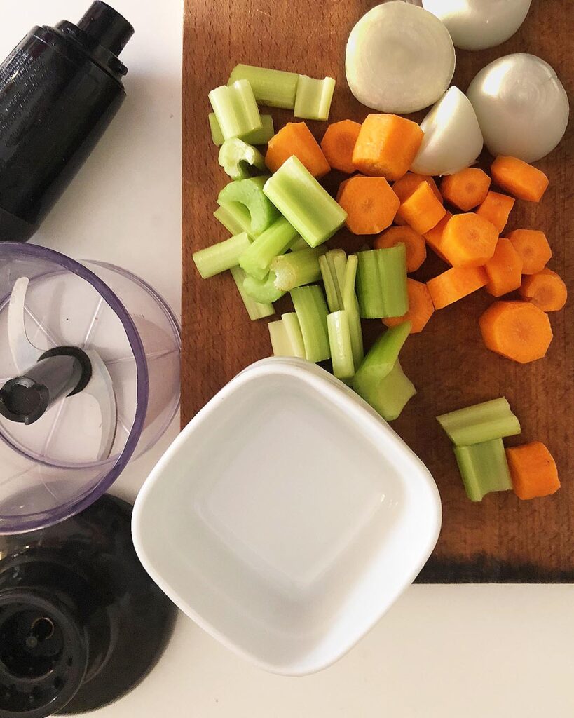 onion, carrot and celery on a chop board with a pan and a blender