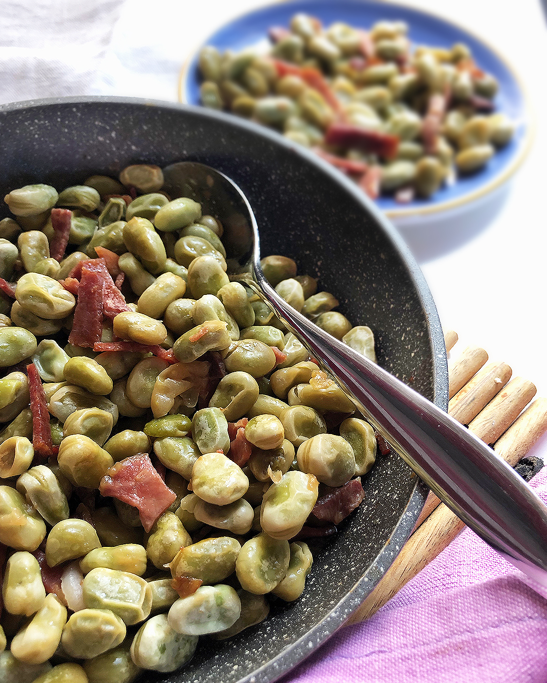 broad beans and prosciutto in a pan with a spoon, other broad beans in the background