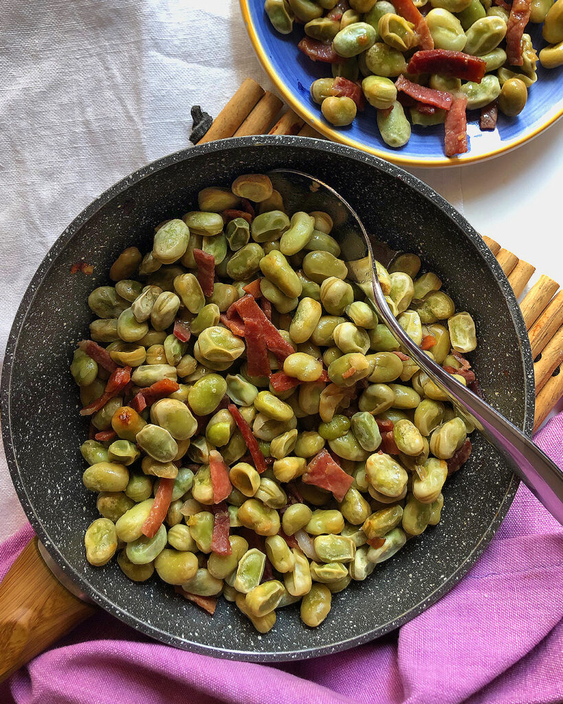 broad beans and prosciutto in a pan with a spoon, other broad beans in the background